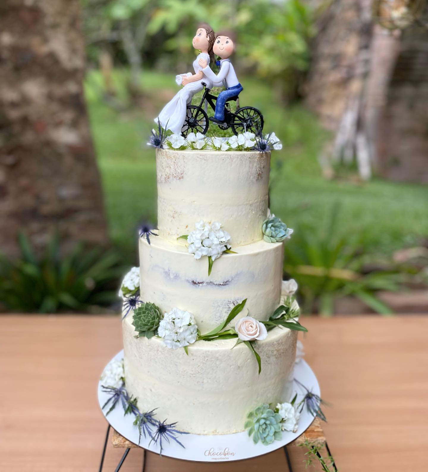 Naked cake de tres pisos con figuras en bicicleta y flores naturales al aire libre