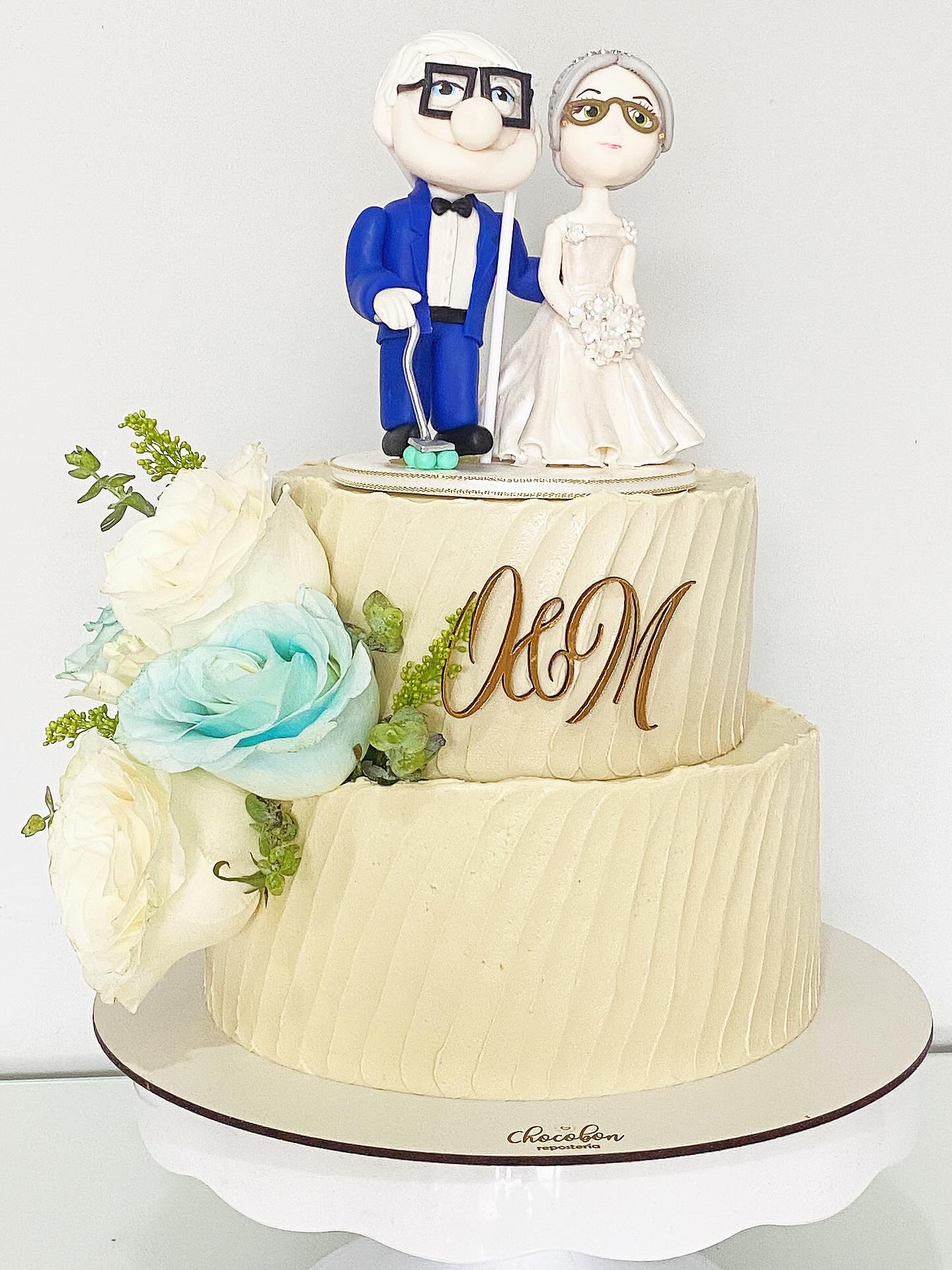 Torta de bodas con figuras de novios mayores estilo Up, monograma dorado J&M y rosas turquesa